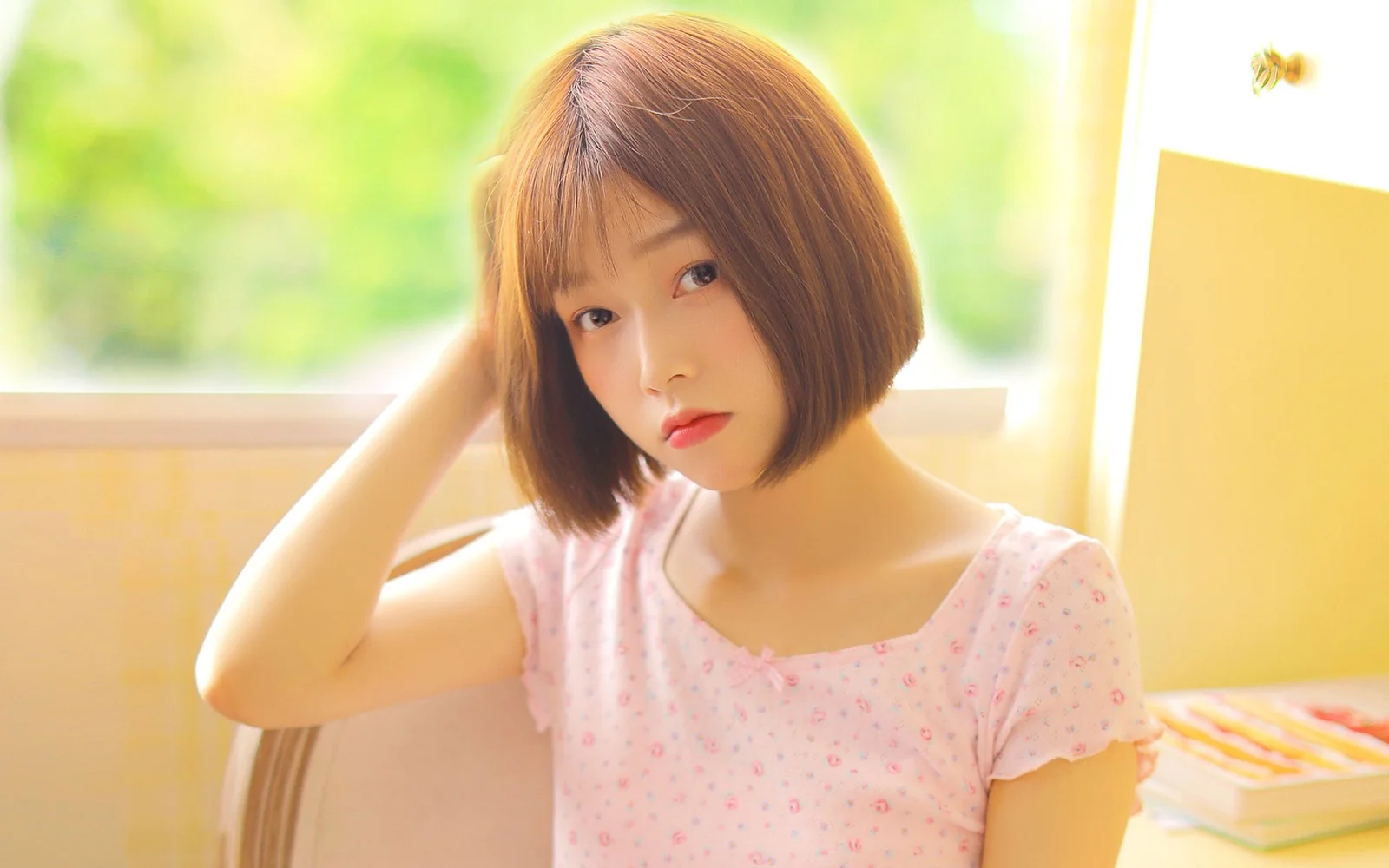 단발머리 여성이 분홍색 원피스를 입고 밝은 창가에 앉아 있습니다.