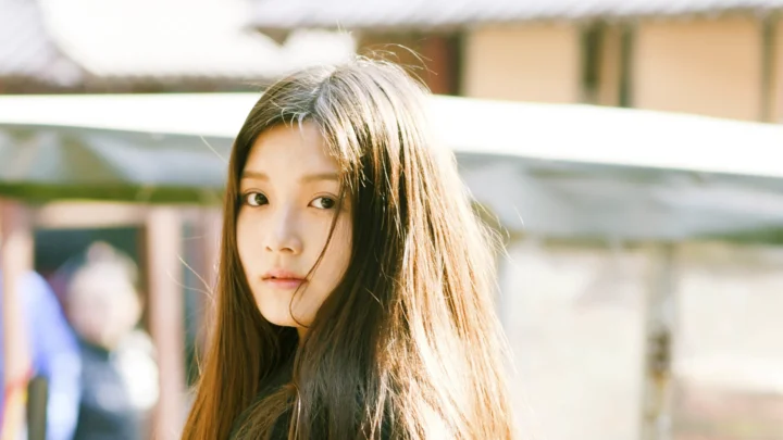 긴 머리의 여성이 야외 햇살 아래에서 카메라를 바라보고 있으며 배경에는 흐릿한 건물이 보인다.