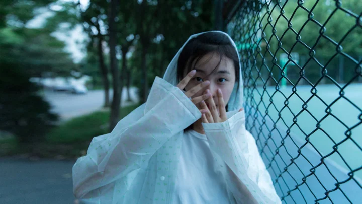 一名身穿白色衣物的女性站在树木和铁丝网围栏旁，用手遮住部分面部。