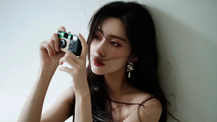 一位成年女性手持相机靠在墙边，黑发被风轻轻拂过。