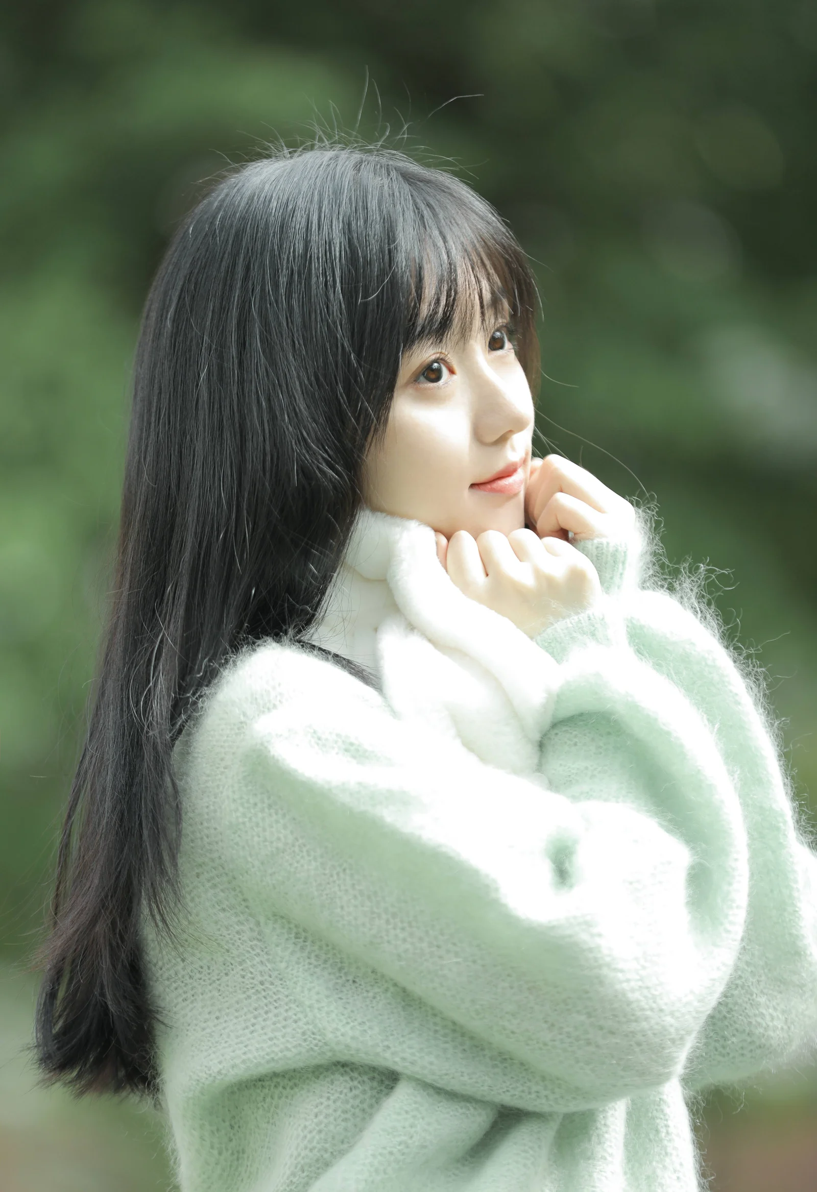 연한 녹색 스웨터를 입은 긴 머리 여성이 흐릿한 녹색 배경 앞에서 손을 턱 가까이에 대고 위를 바라보고 있다.