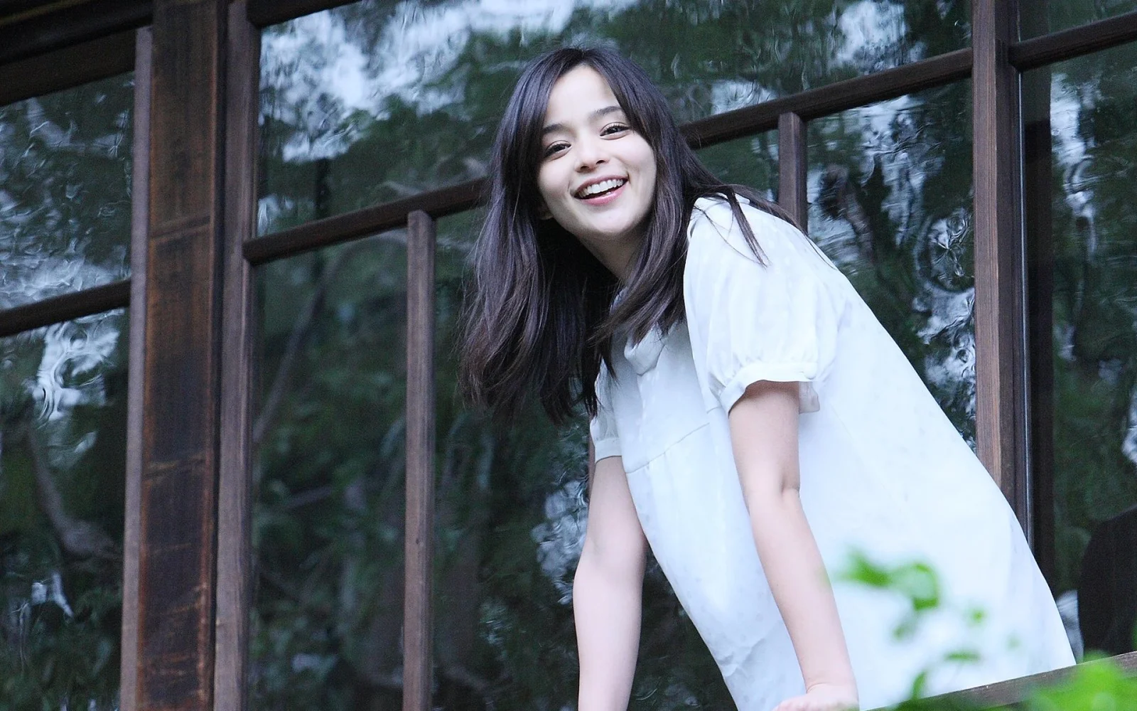 나무 창틀 옆에서 긴 머리의 여성이 미소 짓고 있다.