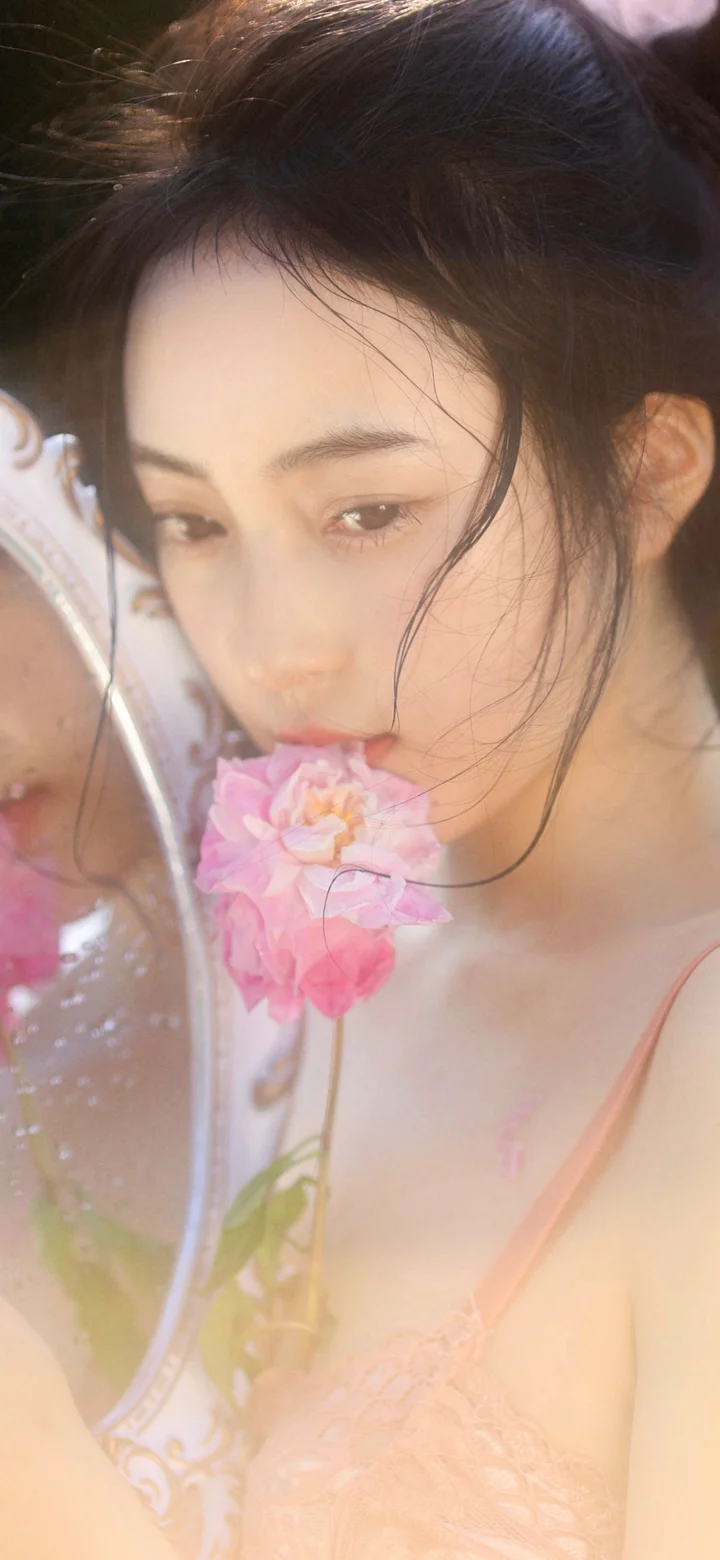一名成年女性在镜前靠近一朵粉色花朵，画面呈现柔和的竖幅人像构图。