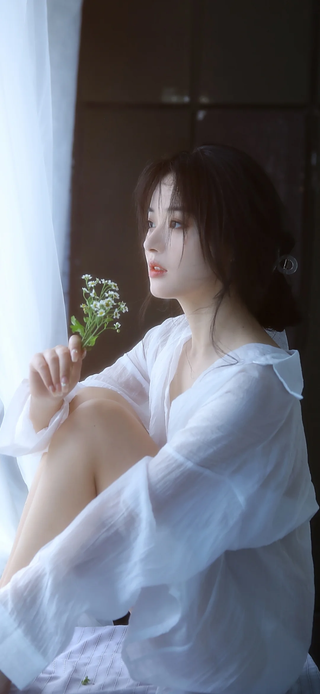 흰 셔츠를 입은 여성이 밝은 창가에 앉아 작은 꽃가지를 들고 있다.