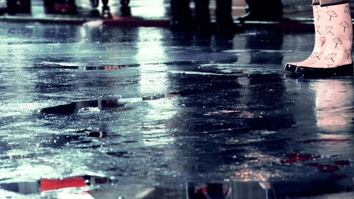 雨水覆盖的深色路面上形成积水和光影倒影，右上角可见一只浅粉色雨靴。