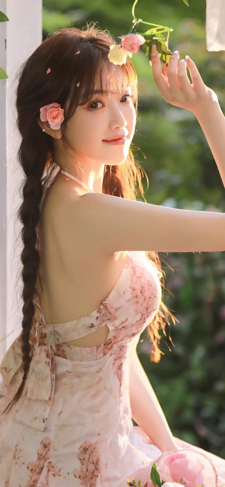 一名成年女子穿着花卉图案连衣裙，在户外阳光下抬手靠近悬挂的花朵。