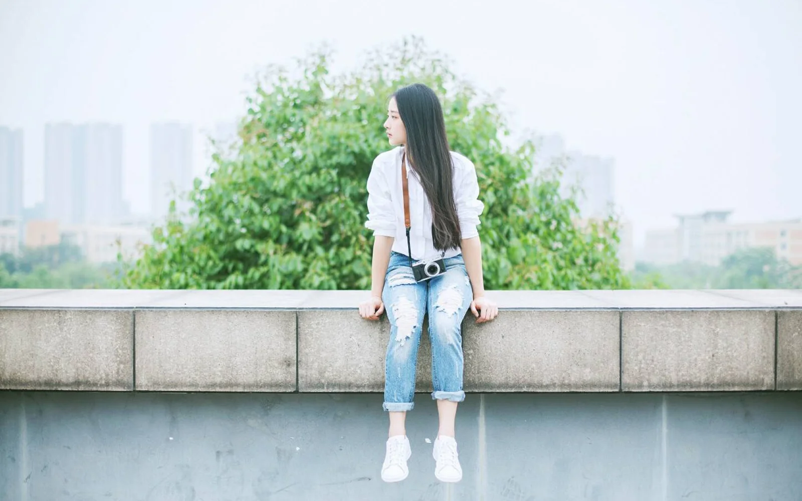 一名女子坐在屋顶边缘，身后是树木和朦胧的城市景观。