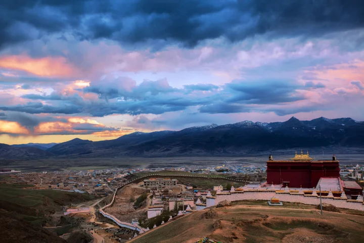 暮色笼罩的高原小镇坐落在群山之间，前景是一座色彩鲜明的寺庙，天空布满绚丽云彩。