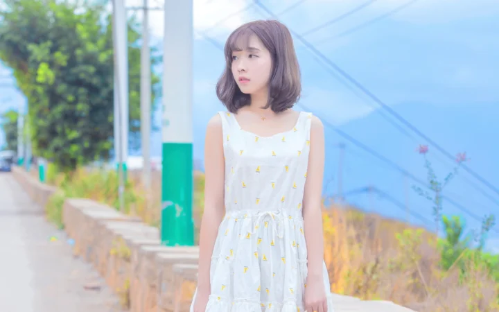 흰색 원피스를 입은 성인 여성이 야외 보도 옆에 서 있다.