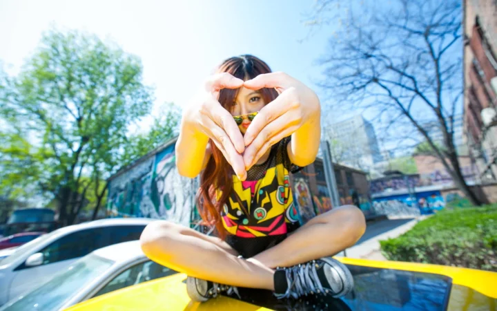 一名女性坐在汽车上，在户外阳光下用双手比出爱心手势。