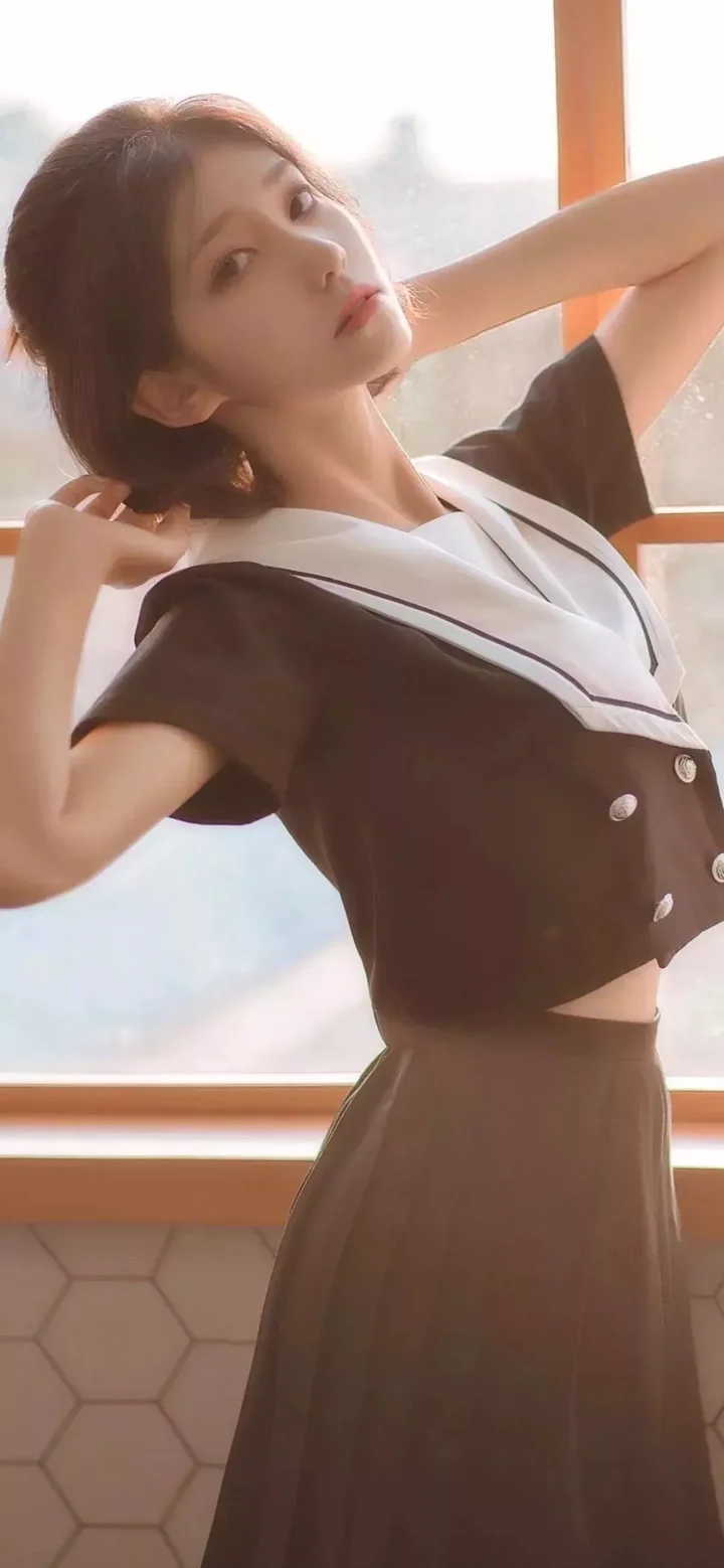 어두운 색 교복을 입은 여성이 창가에 서서 부드러운 빛 속에서 옆모습을 보이고 있다.