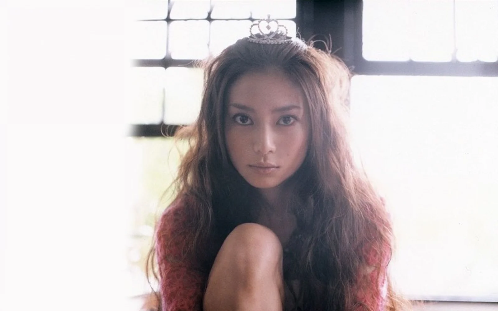 작은 티아라를 쓴 긴 머리 여성이 밝은 창가에 앉아 카메라를 바라보고 있습니다.