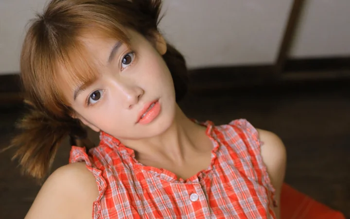 갈색 양갈래 머리의 여성이 빨간 체크무늬 상의를 입고 카메라를 향해 살짝 고개를 기울이고 있습니다.