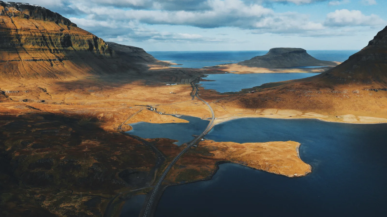 Aerial view of a rugged mountain valley with blue lakes, a coastal fjord, and a winding road.