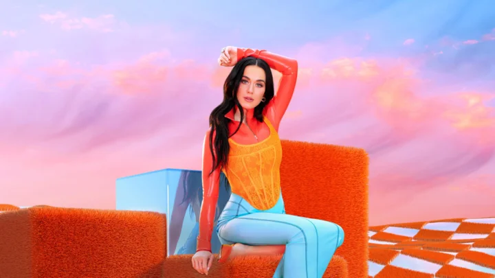 一名黑发女性身穿橙色服装坐在明亮的橙蓝色几何形体之间，背景是粉蓝色天空。