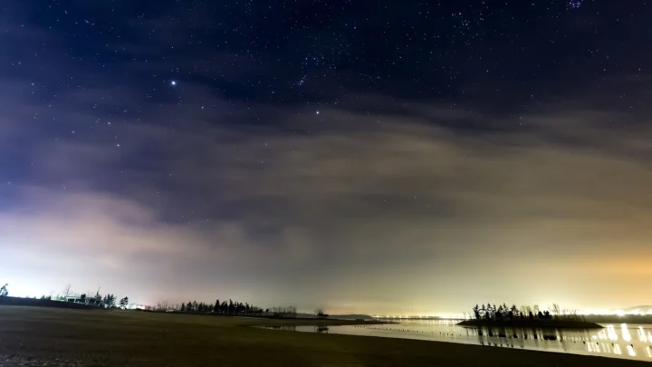 繁星点缀的夜空笼罩着湖畔，云层与远处灯光倒映在平静水面上。