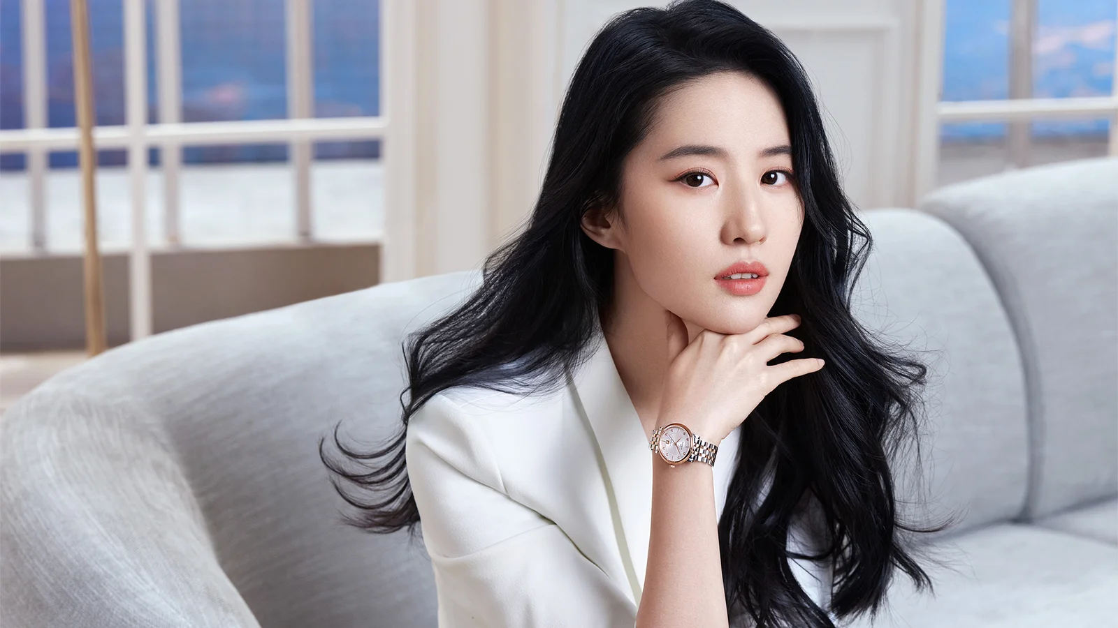 검은 머리의 여성이 흰색 상의를 입고 밝은 창문을 배경으로 연한 색 소파에 앉아 손을 턱 가까이에 대고 있다.