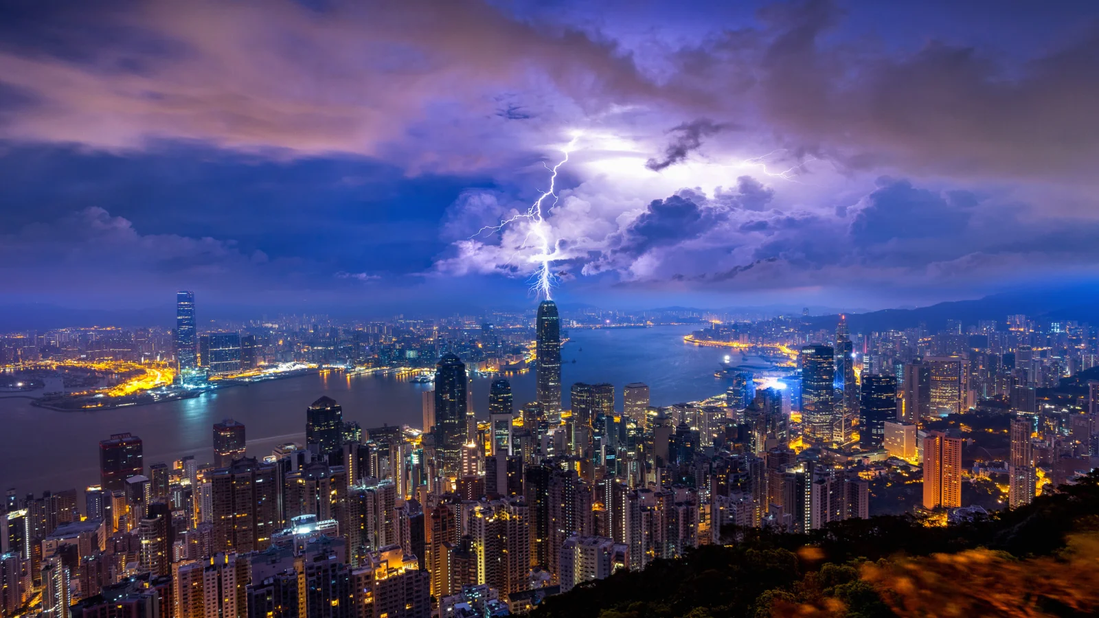 夜の港の両岸に輝く都市の夜景が広がり、嵐の雲の中を明るい稲妻が走っています。