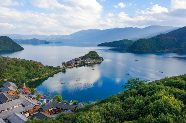 群山环绕的宁静湖泊延伸至远方，前景可见森林、村落屋顶和水面上的小船。