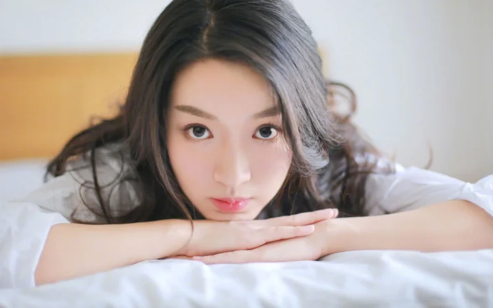 一名成年女性俯卧在白色床铺上，将下巴 resting 在交叠的手臂上看向镜头。