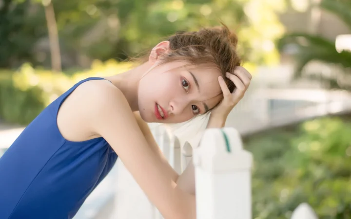 一名身穿蓝色连衣裙的女子在绿意环绕的户外靠着白色栅栏凝视镜头。