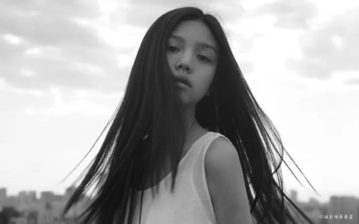 一名长发女性站在户外天空与城市背景前的黑白肖像画面。