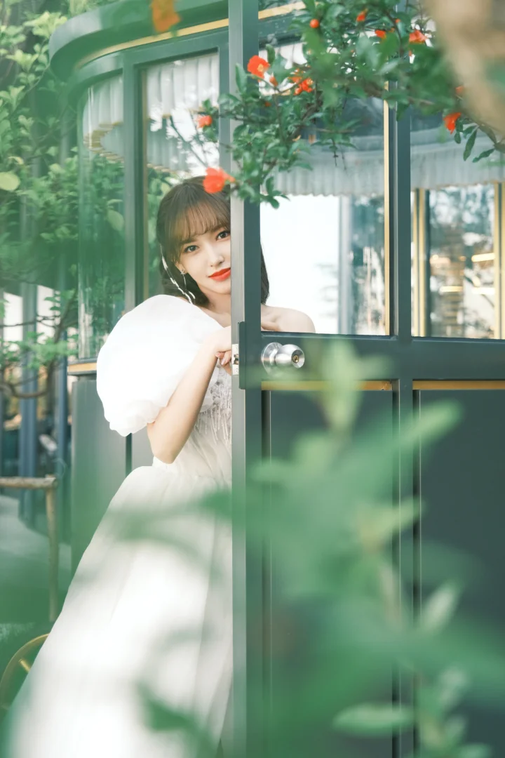 一位穿白色服装的成年女性从绿门后探出身来，周围有绿植和橙色花朵。