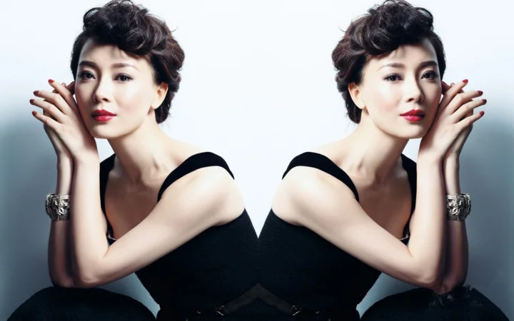 画面中一位身穿黑色连衣裙的成年女性以镜像构图面对镜头，背景为浅灰色。