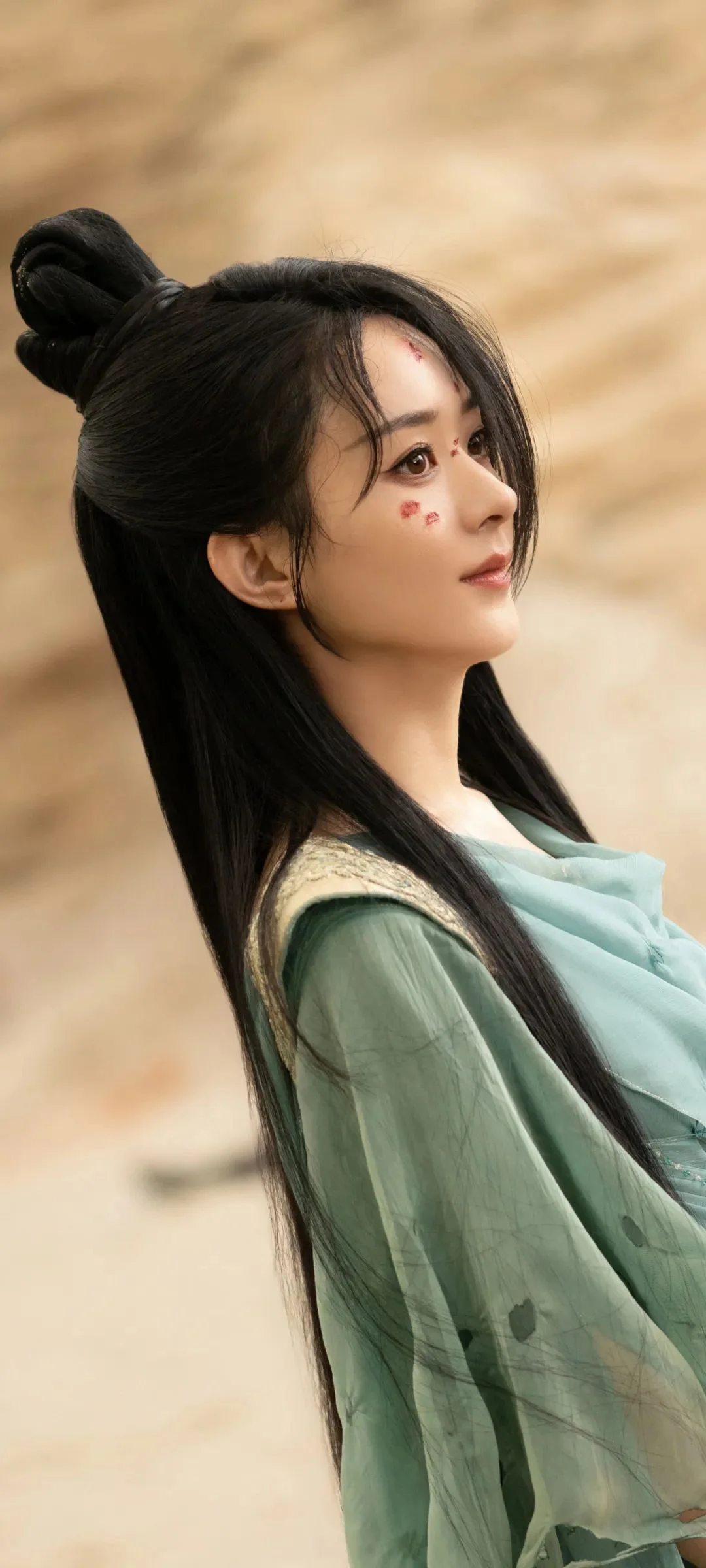 초록색 의상을 입고 긴 머리를 한 여성이 따뜻한 베이지색 배경을 뒤로하고 옆모습을 보입니다.
