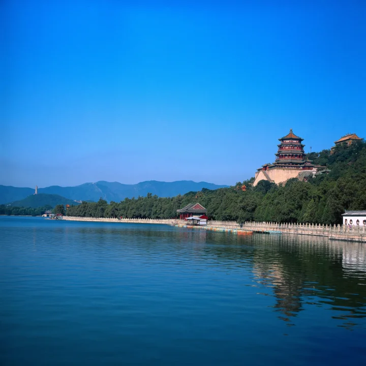 宁静湖面映照着湖畔的中式古建筑、葱郁树木与远处群山，天空湛蓝。