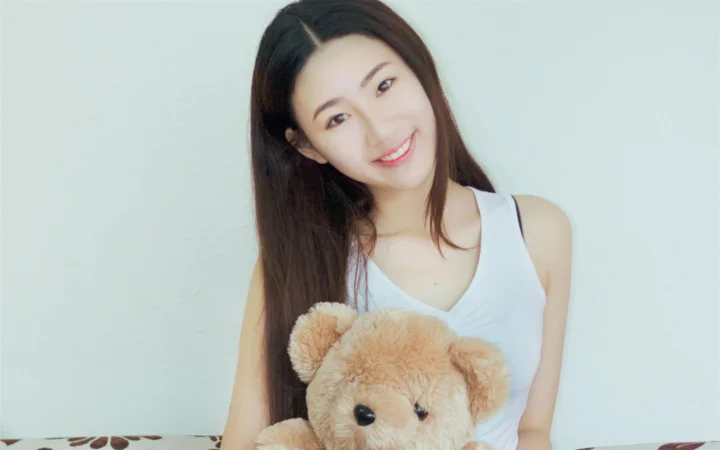 긴 머리의 여성이 밝은 색 벽을 배경으로 갈색 테디베어를 안고 미소 짓고 있다.