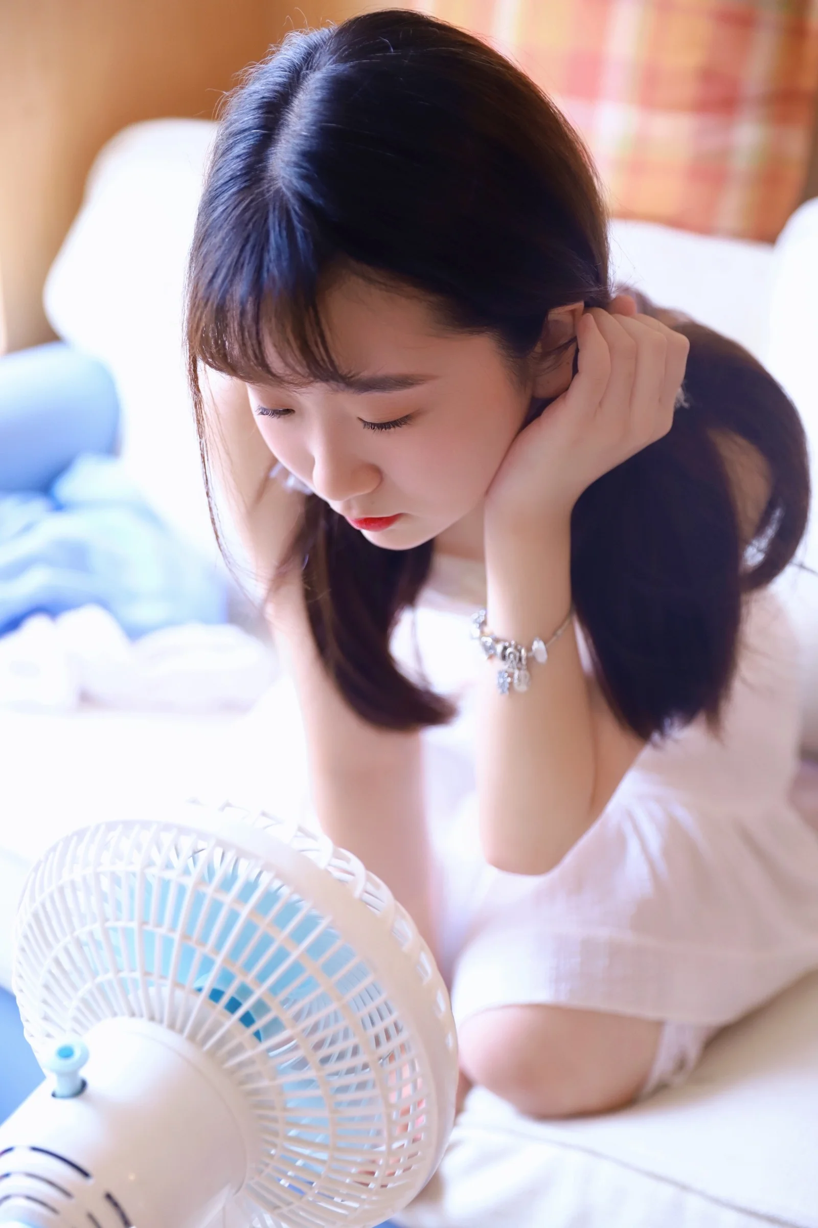 검은 머리의 여성이 실내 침대 옆에 앉아 귀 근처에 손을 대고 있으며 앞쪽에는 흰색 선풍기가 놓여 있습니다.