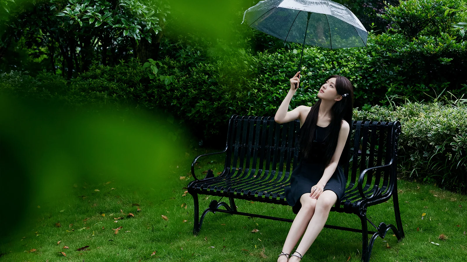 一名身穿黑色衣服的女子坐在花园长椅上，手持透明雨伞，周围是茂密的绿色植物。