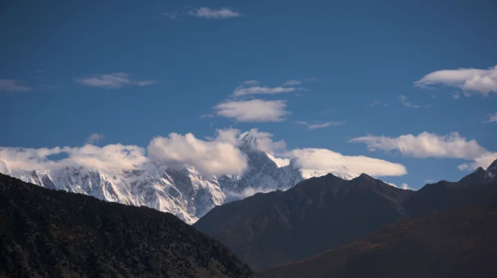 蓝天下，云雾环绕的雪山与前景山脉构成壮阔的自然风景。