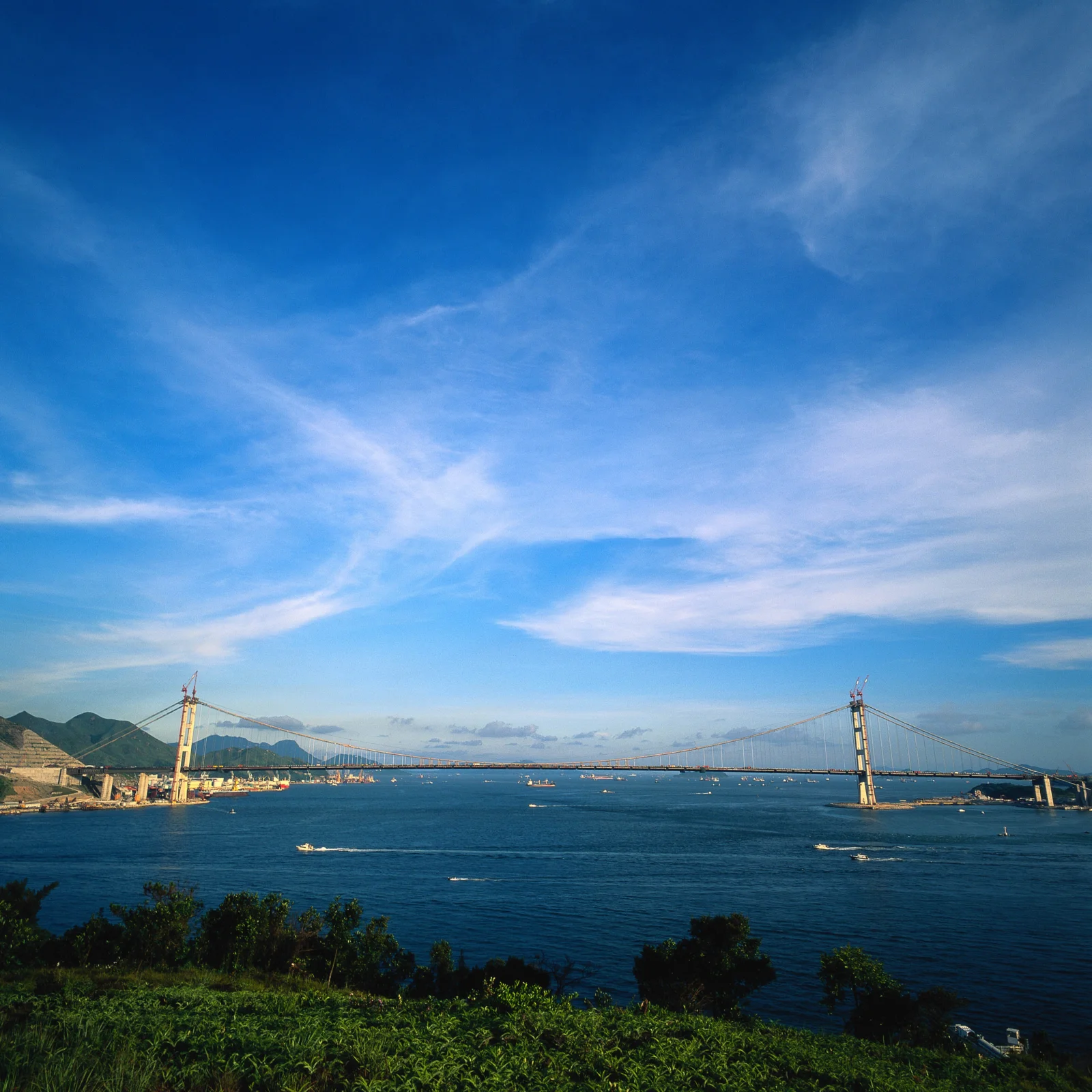一座悬索桥横跨蓝色海湾，远处是群山，天空布满淡淡的云彩。