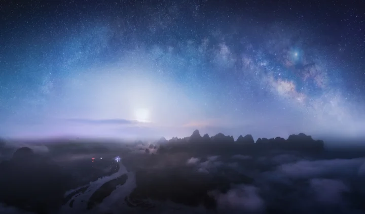 繁星密布的银河横跨夜空，雾气笼罩的山脉在下方形成深色剪影。