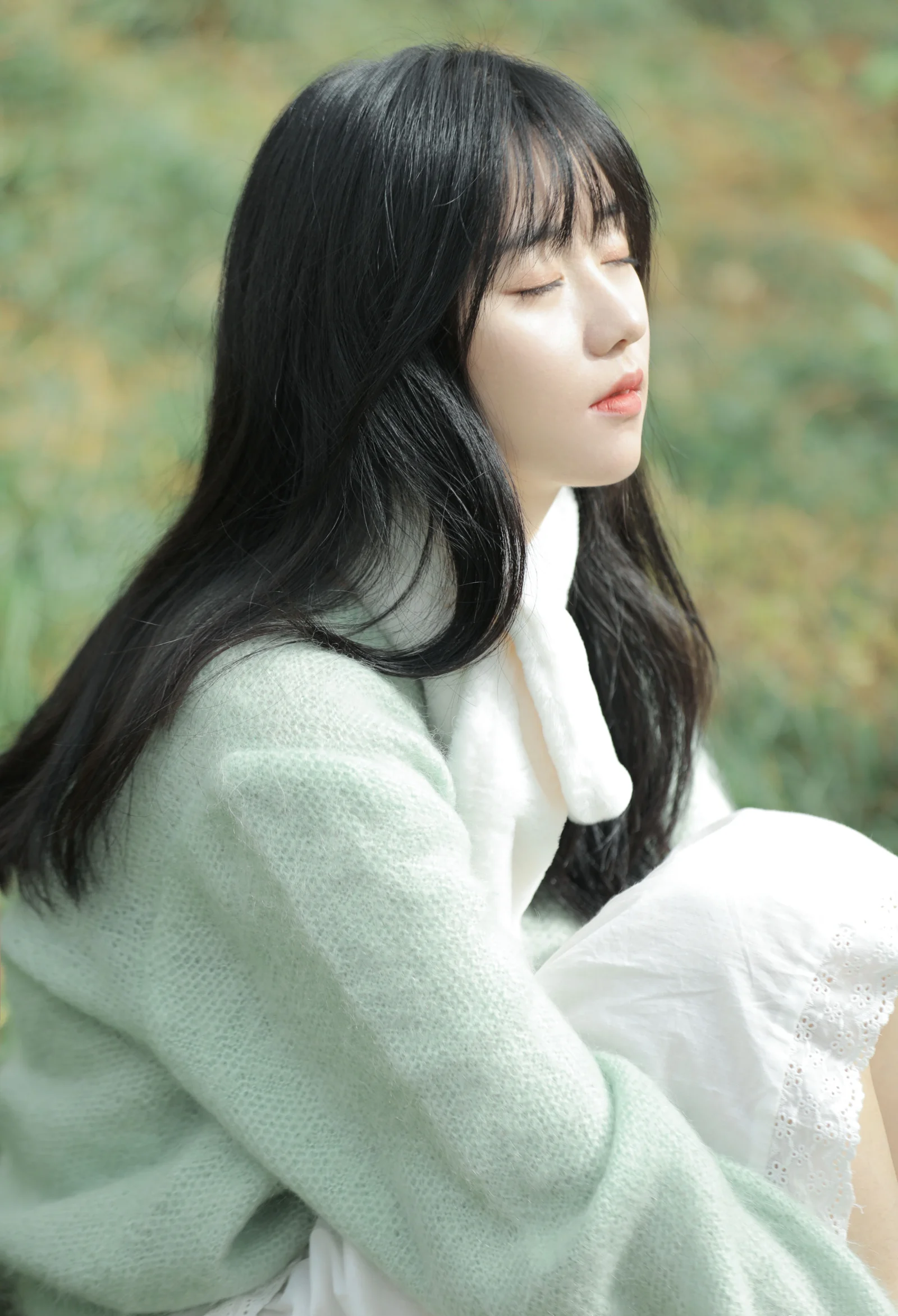 긴 머리의 여성이 밝은 색 스웨터를 입고 야외에 앉아 눈을 감은 옆모습을 보입니다.