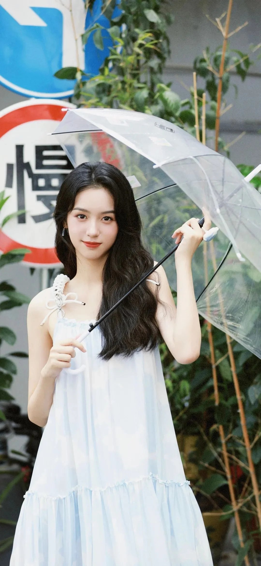 一名长发女子穿着白色连衣裙，在绿植和标牌前撑着透明雨伞。