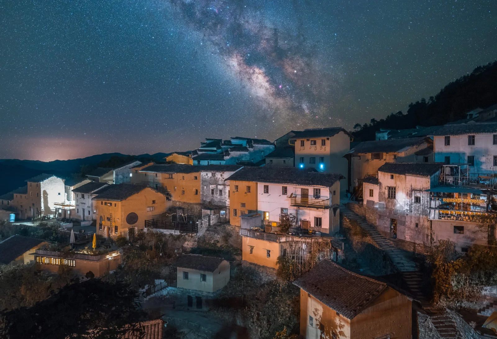 夜空中的银河横跨山村上方，照亮了层叠的房屋与远处的山峦。