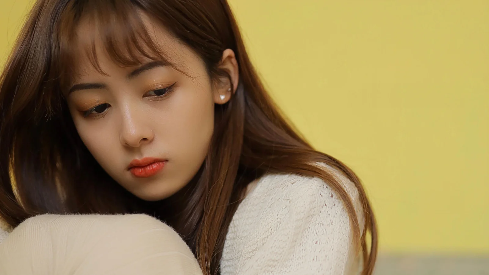 연한 색 니트 스웨터를 입은 갈색 머리 여성이 부드러운 노란색 배경 앞에서 무릎 곁에 기대어 아래를 바라보고 있습니다.