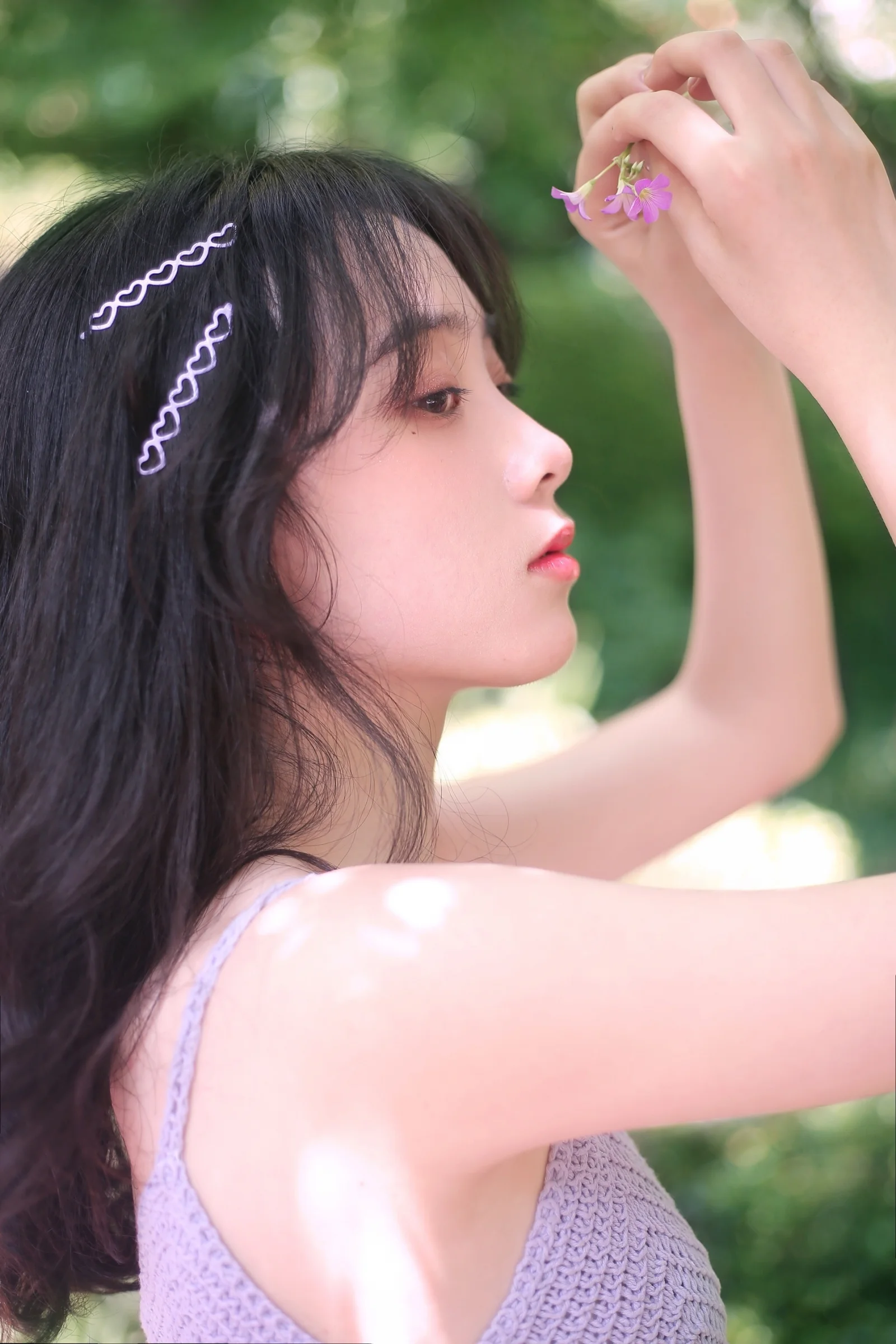 一位长发女性在户外绿意背景前举手靠近紫色小花，呈现柔和的侧脸光影。