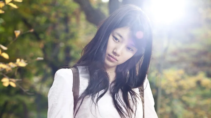 一名长发女子身穿白色上衣，在林间柔和阳光下站立。
