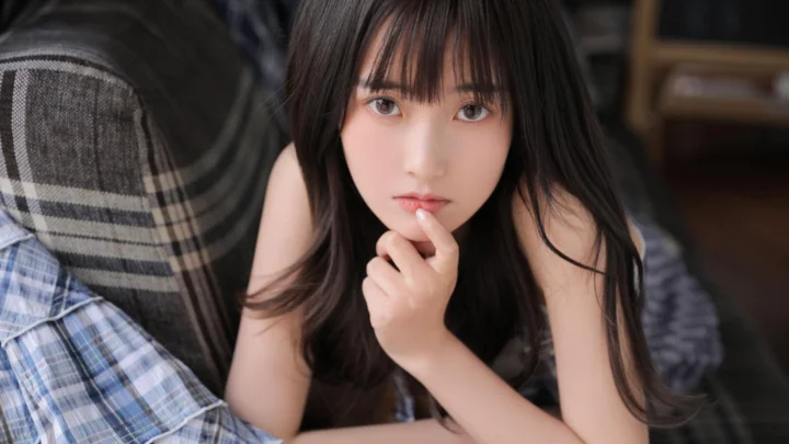室内で長い髪の女性がカメラを見つめ、指先をあごに添えています。