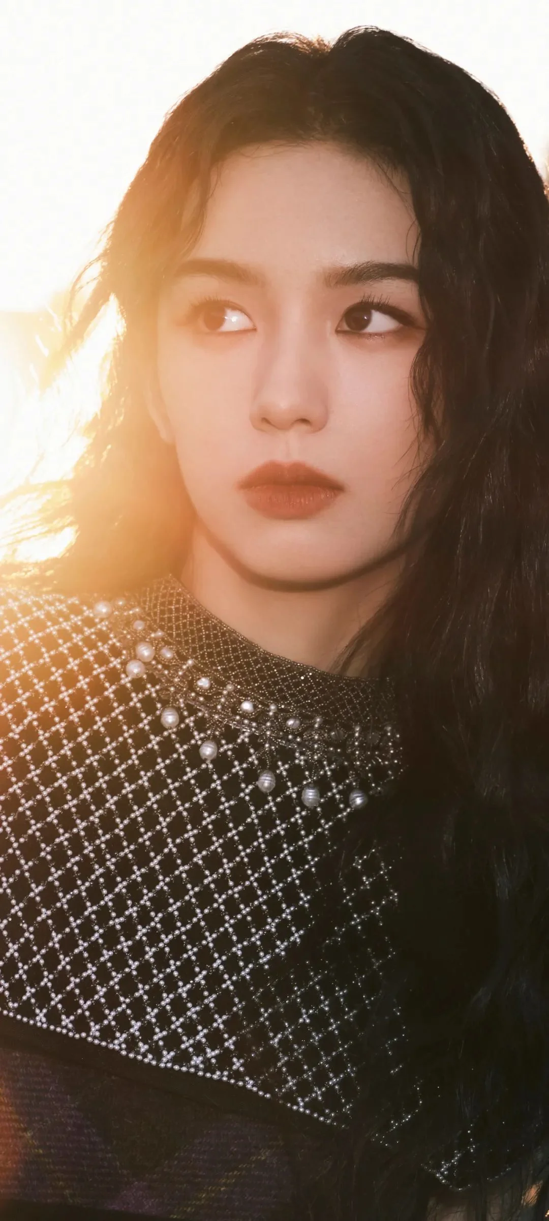 一位黑发女性在暖色逆光中侧目凝视，身穿饰有珠光网格的深色上衣。