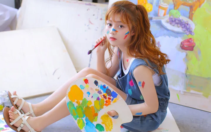一名年轻女子坐在画室中，手持画笔并拿着调色板，周围可见画布和彩色颜料。
