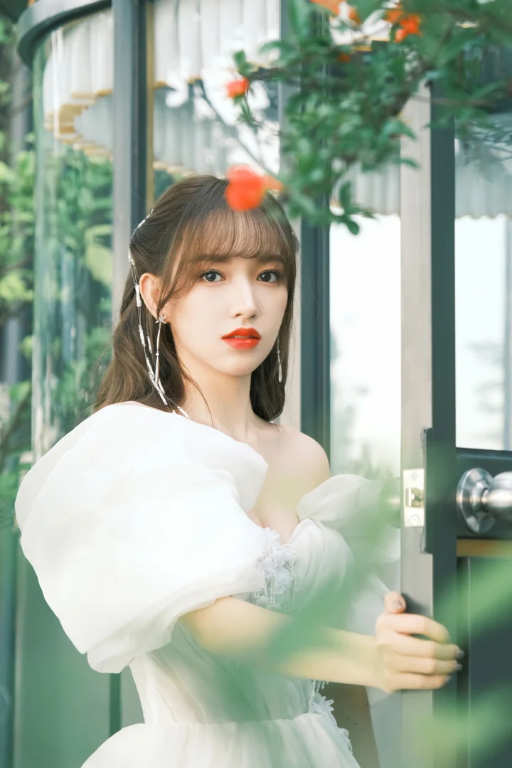一名成年女子穿着白色连衣裙站在玻璃门旁，周围可见绿植与柔和光线。
