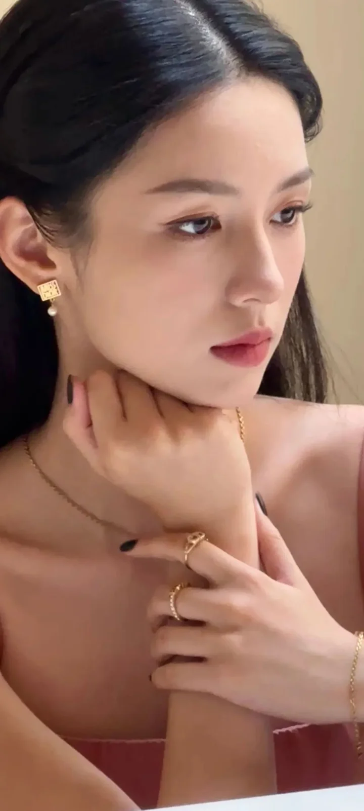 一名女性在柔和室内光线下侧脸凝视，手托着颈部并佩戴金色耳饰、项链和戒指。
