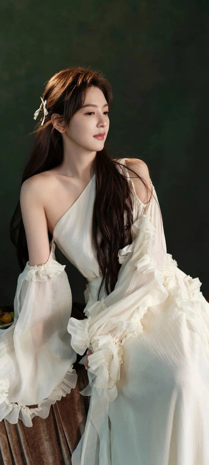 一名長髮女子身穿白色連衣裙坐在深色背景前。