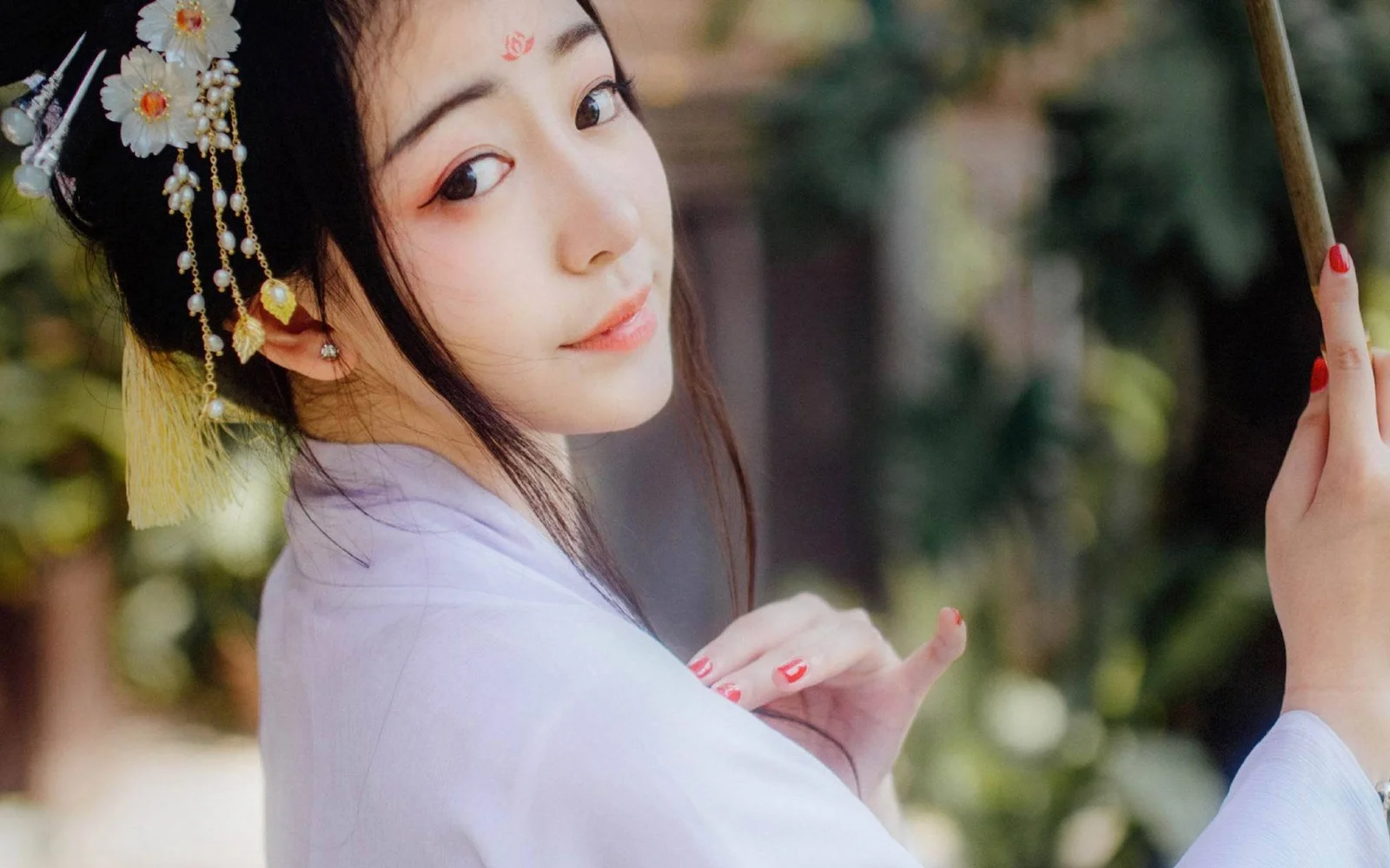 一位佩戴花饰与流苏发饰的女子在户外回望镜头，背景为虚化的绿色植物。