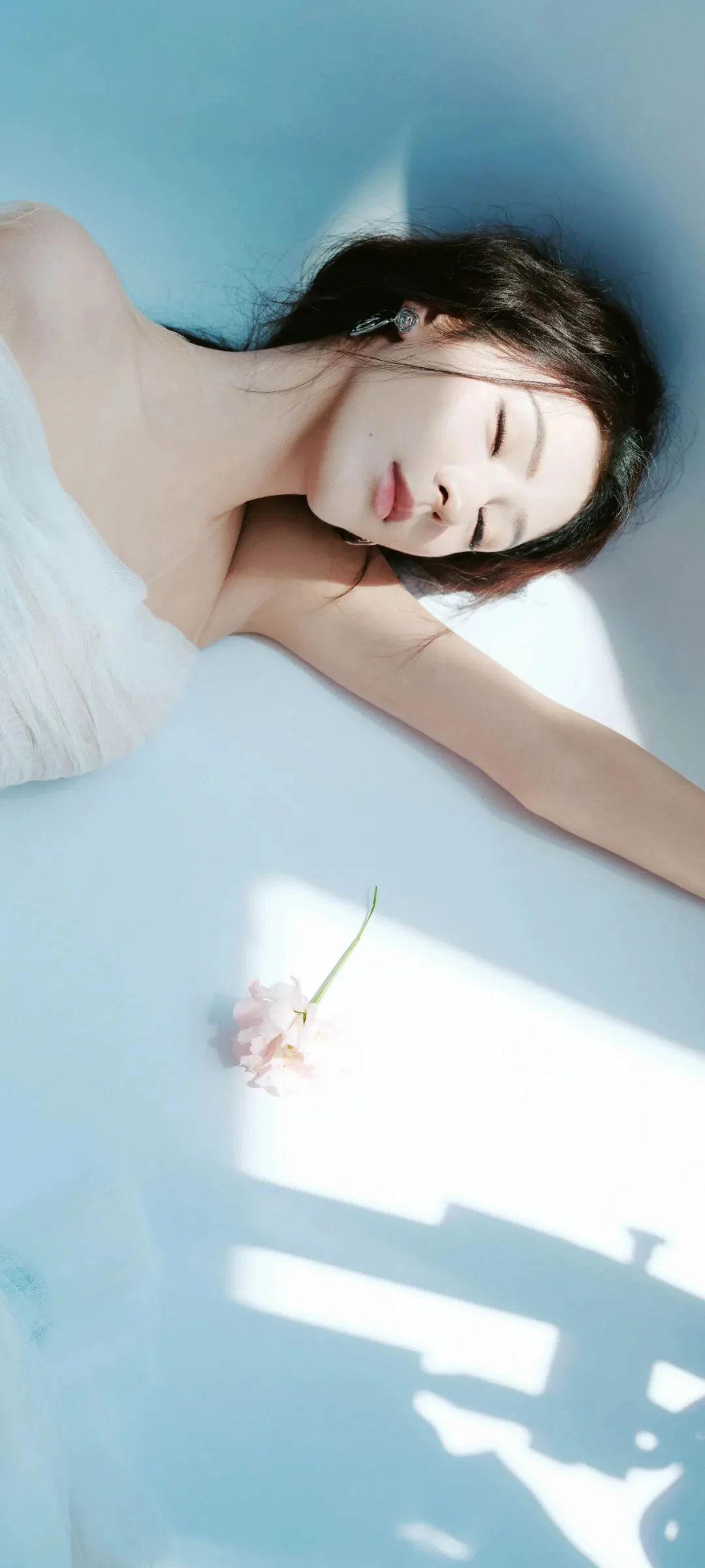 目を閉じた女性が白い服を着て淡い青色の背景に横たわり、周囲に日差しと窓の影が映っています。